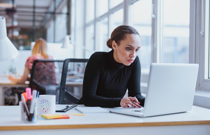 woman in black turtle neck intently staring at laptop at desk in office