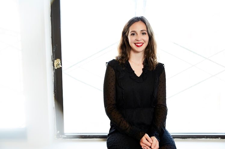 A brunette woman smiling a black dress as she is leaning next to a window