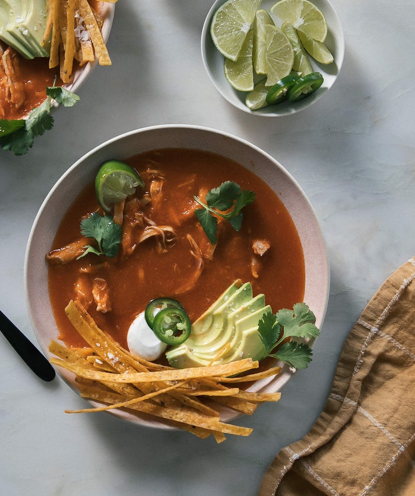 bowl of red soup garnished with sliced avocado, tortilla chips, cilantro, jalapeno peppers, and lime...