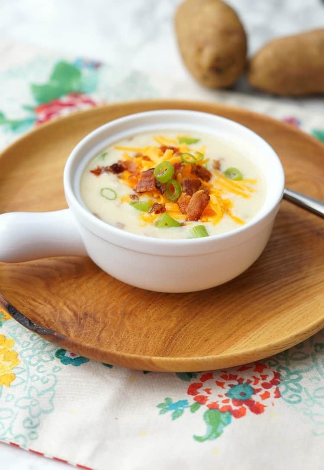 white bowl of white potato soup garnished in shredded cheddar cheese, bacon and chives on top of a w...
