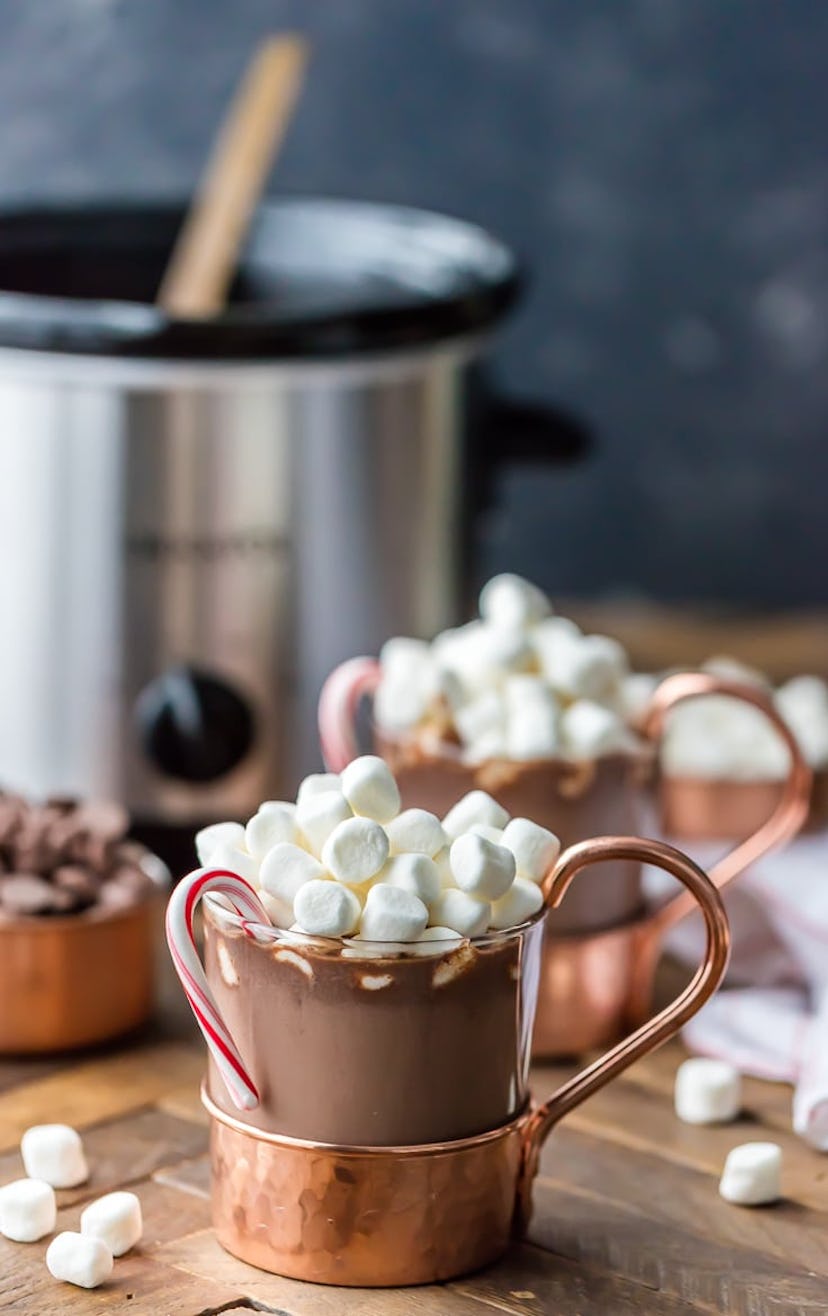 Copper and glass mugs of hot chocolate with marshmallows on top and a candy cane hanging off the lef...