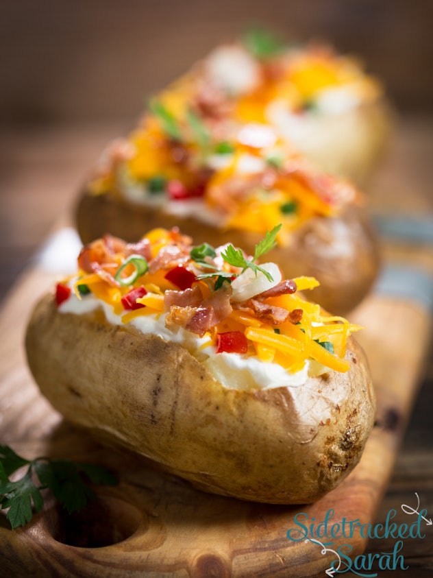 close up of baked potatoes garnished with shredded cheese, bacon and chives on a wooden cutting boar...