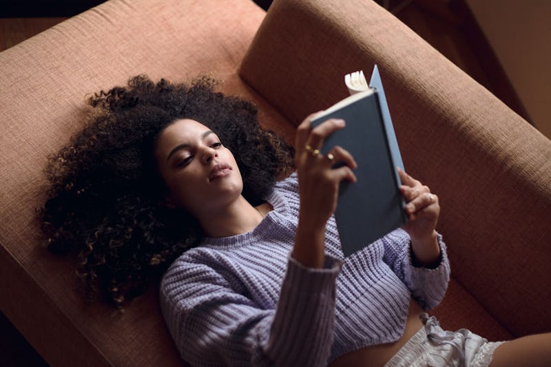 A woman in a blue sweater lying on a brown couch reading a book
