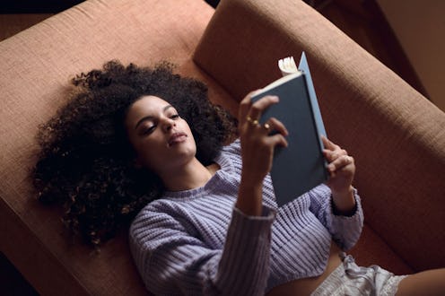 A woman in a blue sweater lying on a brown couch reading a book