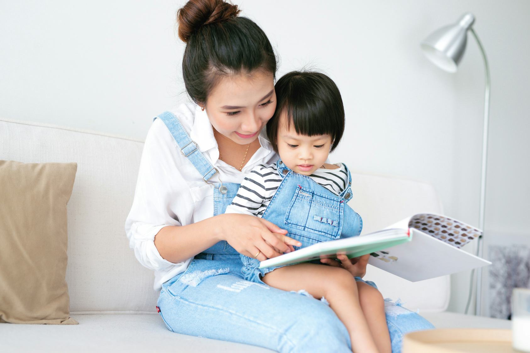 A mother in her 20s holding her daughter in her lap while they read a book.