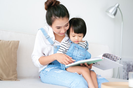 A mother in her 20s holding her daughter in her lap while they read a book.