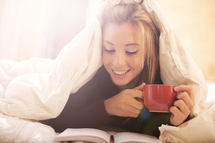 A girl reading a book and drinking coffee while lying in the bed covered with sheets.