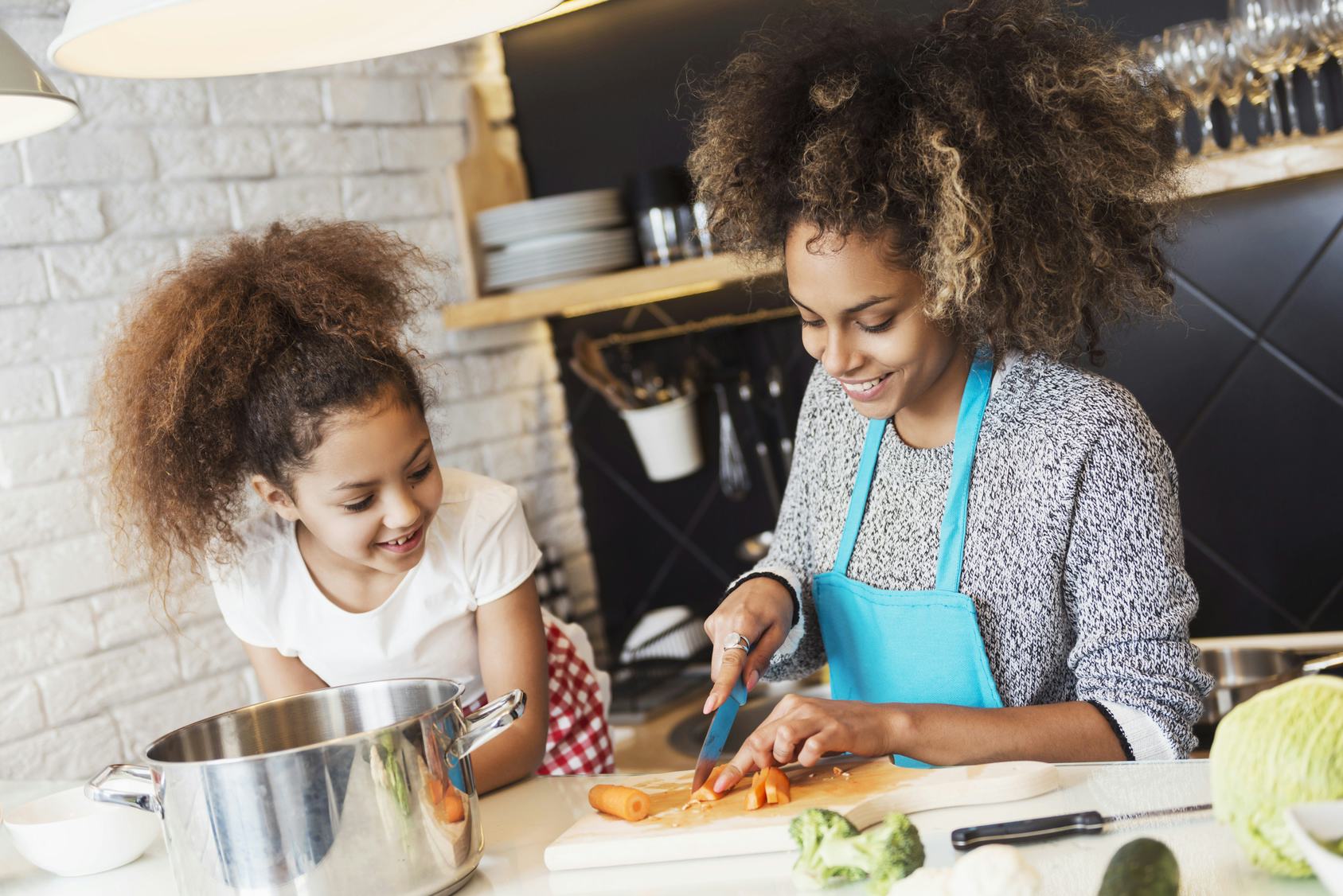 A woman wearing an apron cutting up carrots while her daughter stands next to her and helps her cook