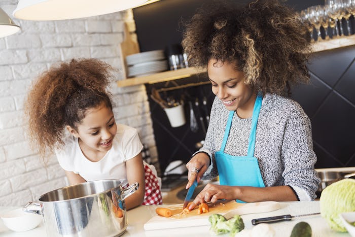 A woman wearing an apron cutting up carrots while her daughter stands next to her and helps her cook