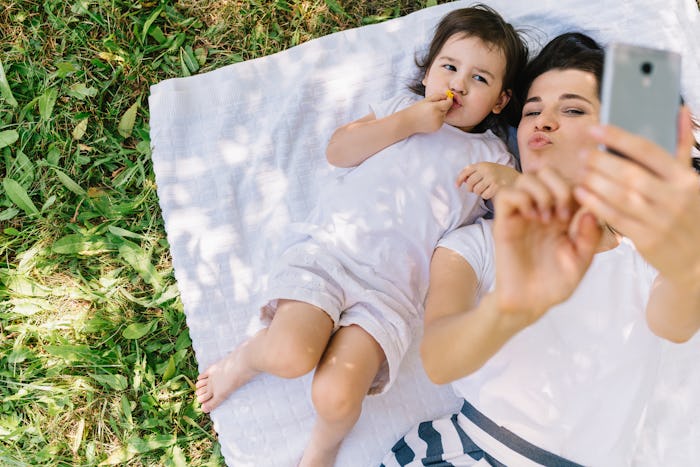 Mom and daughter both in white lying on a blanket on the grass taking a selfie