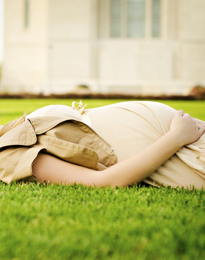 A pregnant woman in a beige dress and jacket lying on her back on green grass