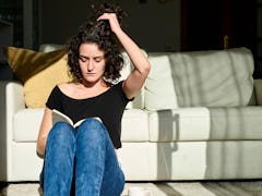 A curly-haired woman sitting on the floor reading a book while holding her hair up.