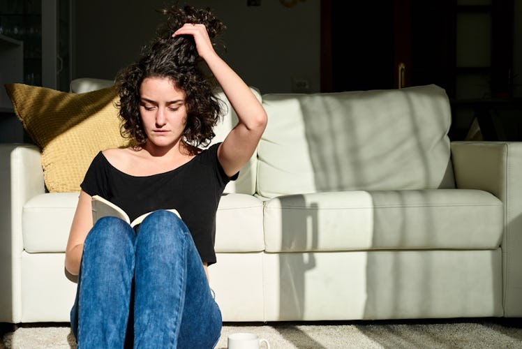 A curly-haired woman sitting on the floor reading a book while holding her hair up.