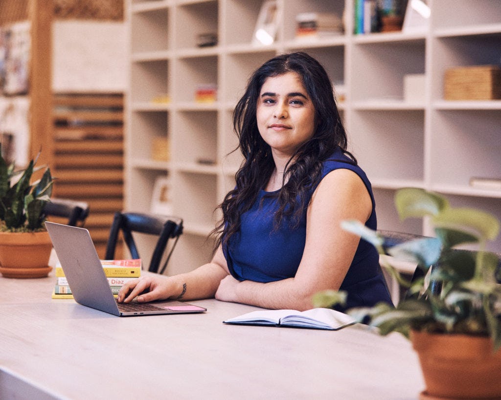 A dark-haired woman in a blue top sitting in her office typing on her laptop