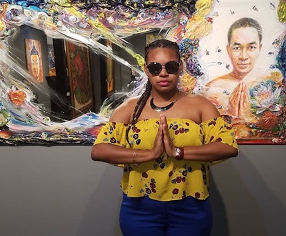 Francis Carrero in a yellow top with her hands clasped together in front of a painting doing the sam...