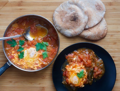 Shakshuka eggs in a pan and served for one person on a black plate next to three pieces of flat brea...