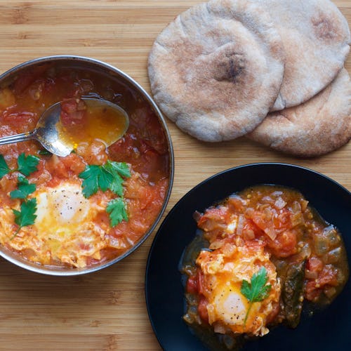 Shakshuka eggs in a pan and served for one person on a black plate next to three pieces of flat brea...