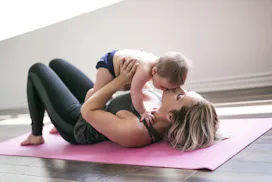 A mom lying on a yoga mat holding her newborn above her and kissing its forehead
