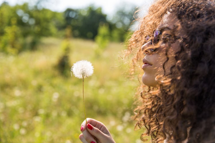 A curly-haired woman with a splitting headache blowing into a dandelion in a park
