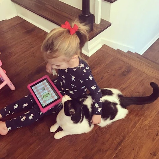 Little girl playing on her tablet while petting cat sitting beside her