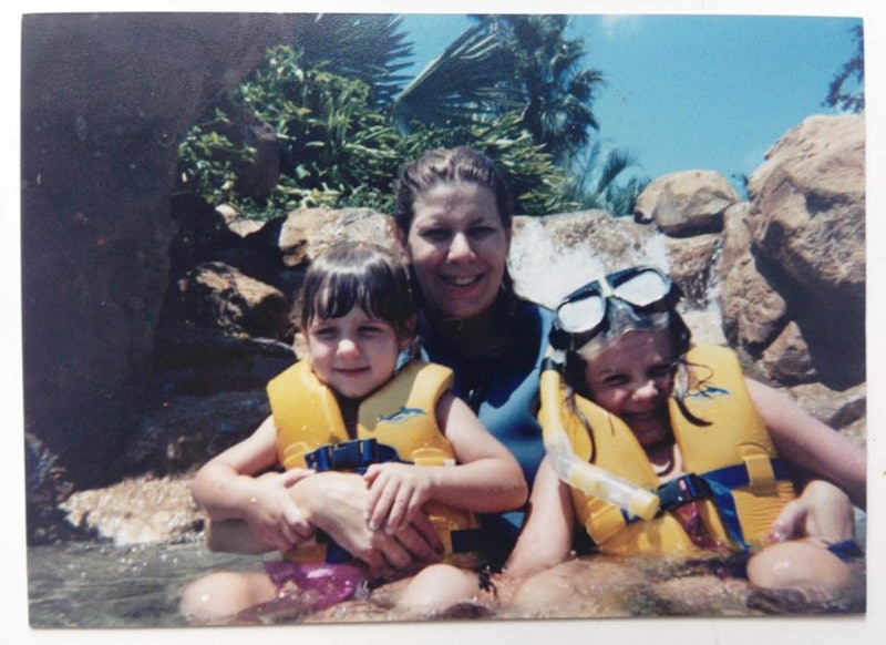 A mom sitting in the water with her two kids, both of them are wearing yellow life jackets