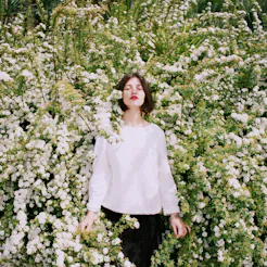 A woman suffering from a hay fever standing in a middle of a flower bush with her eyes closed