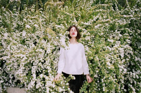 A woman suffering from a hay fever standing in a middle of a flower bush with her eyes closed