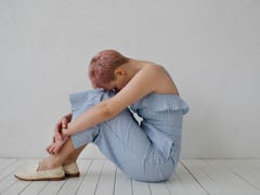 A woman with pink hair, wearing a blue romper sitting on the floor curled up due to period cramps