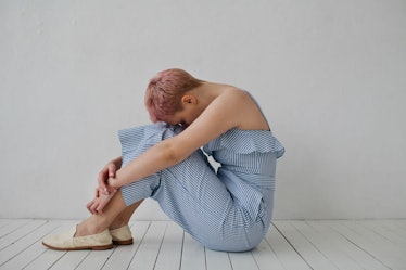 A woman with pink hair, wearing a blue romper sitting on the floor curled up due to period cramps