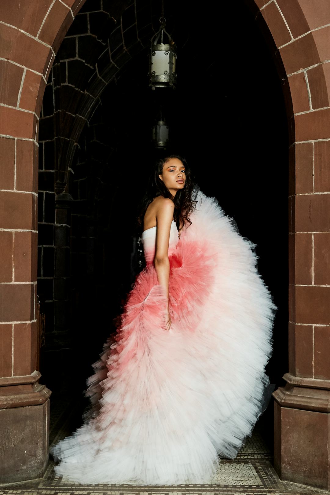 A woman standing at the beginning of an alleyway wearing a puffy white and pink dress