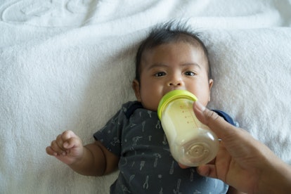 A baby laying in the bed and drinking milk from the bottle his mom is holding
