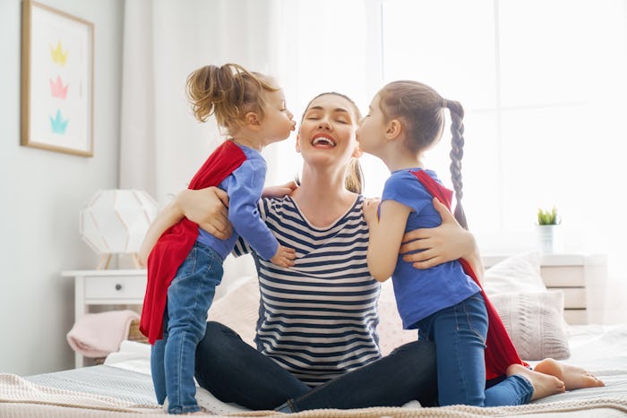 A mother being kissed by her daughters who are wearing red superhero capes on Mother's Day