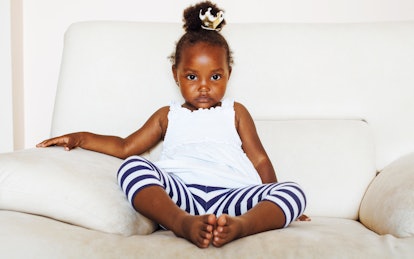 A little girl is sitting on a white couch wearing a white shirt and blue striped leggings with a bun...