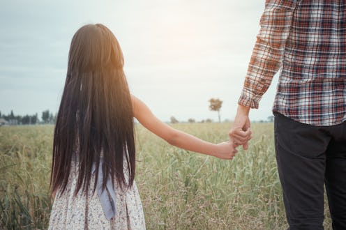 A girl with a long brown hair, wearing a white dress while holding her father's hand in a field
