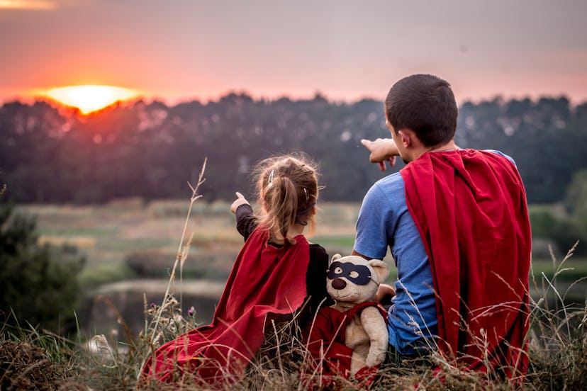 A father, daughter and her teddy bear wearing matching superhero costumes while watching the sunset