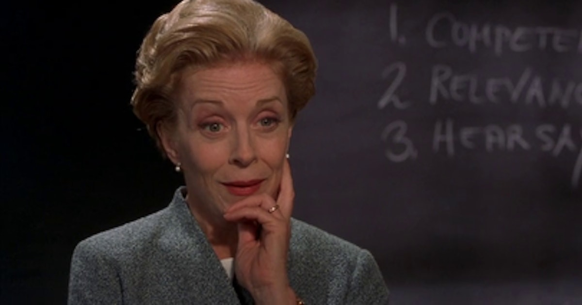 Still from Legally Blonde. A close up of Professor Stromwell, an older woman and law professor standing in front of a chalk board in a grey suit. She is holding her chin with an amused expression.