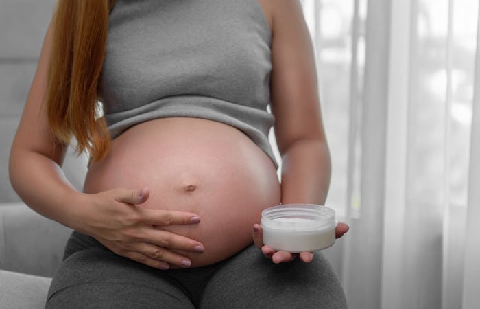 close up shot of pregnant woman bare belly one hand against belly the other holding jar of white cre...