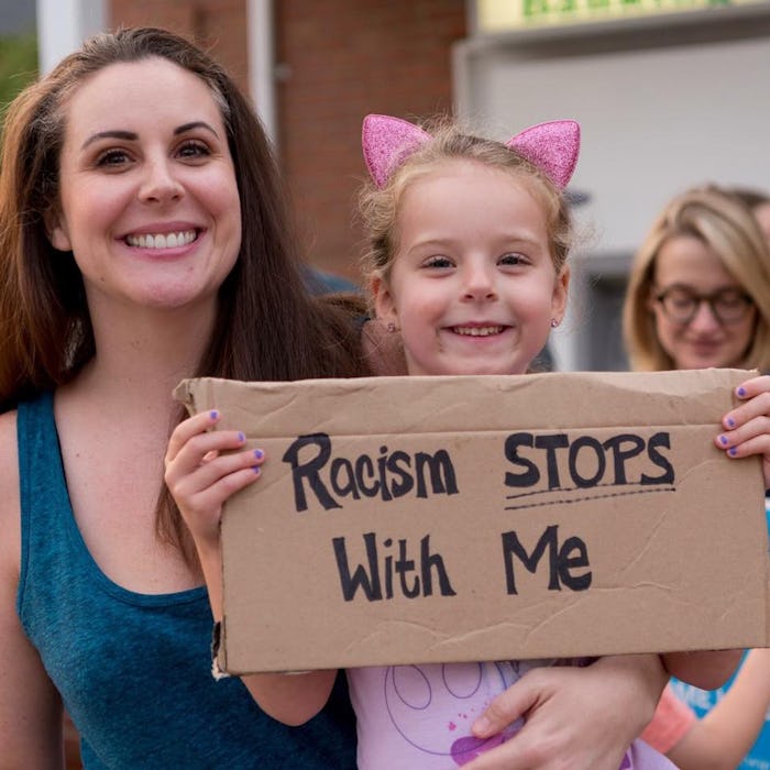 A little girl holding a cartoon banner with "racism stops with me" text while standing next to her m...