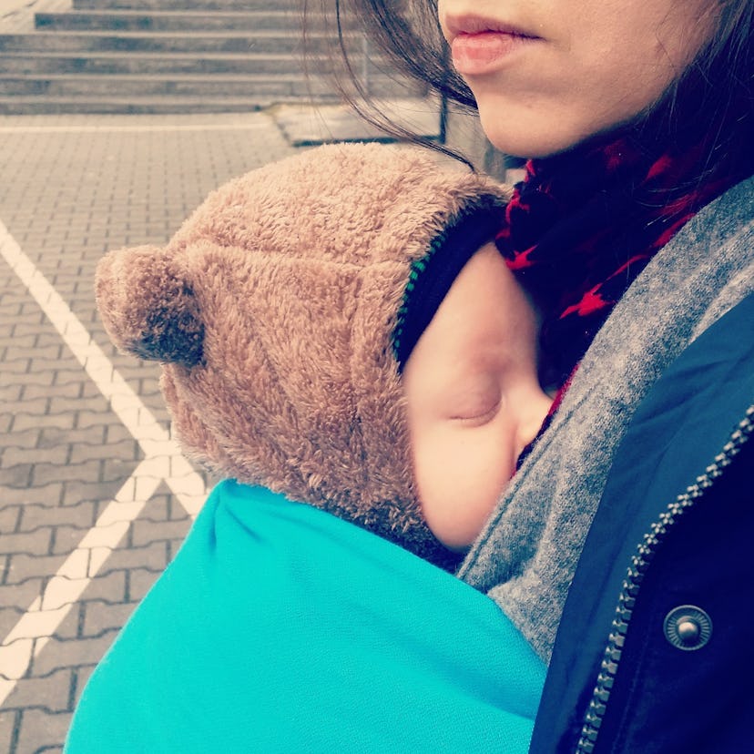 Woman carrying a sleeping baby wearing a brown teddy bear hat in a blue sling
