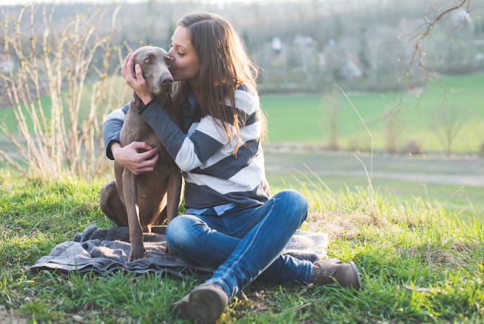 A woman hugging and kissing her dog while sitting on the grass with a river in the background.