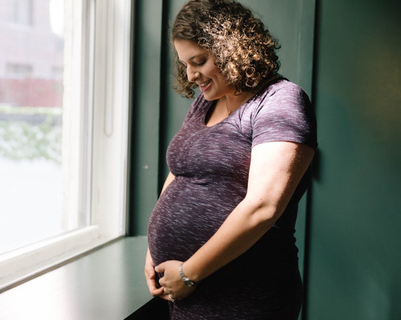 pregnant woman with curly brown hair holding round belly next to window smiling