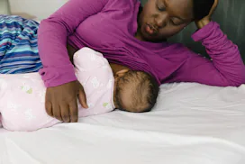 Woman in purple shirt breastfeeding her baby on their bed