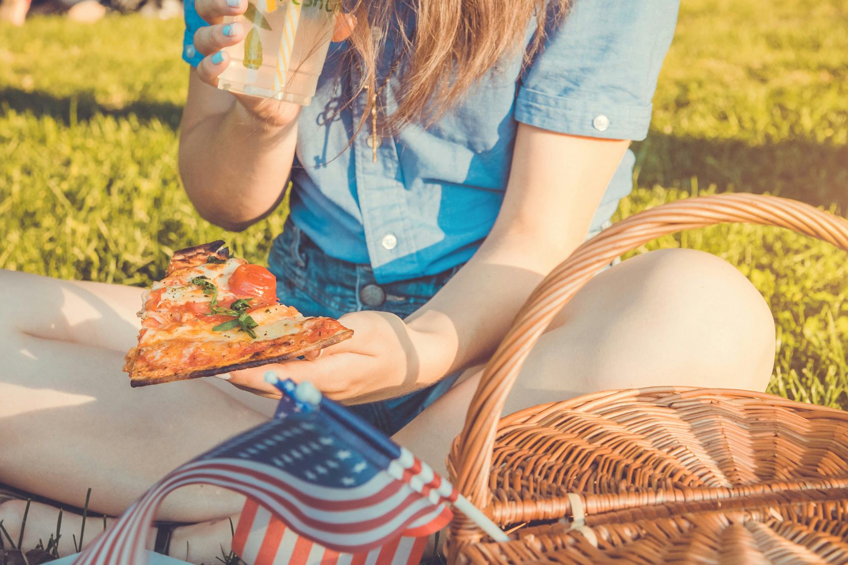 A woman sitting on the grass with her picnic basket next to her, eating pizza