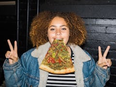 A curly girl holding a slice of pizza in her mouth while showing a piece sign with both of her hands...