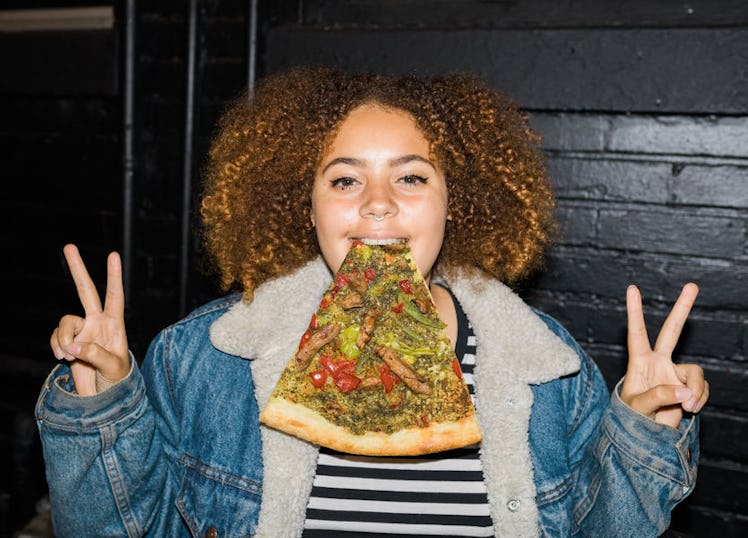 A curly girl holding a slice of pizza in her mouth while showing a piece sign with both of her hands...