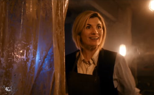 Jodie Whittaker as the doctor, standing in a poorly lit room smiling and looking off to the side