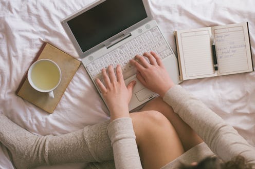 A woman on a bed with a cup of tea, and a notebook in front of her, typing on her laptop