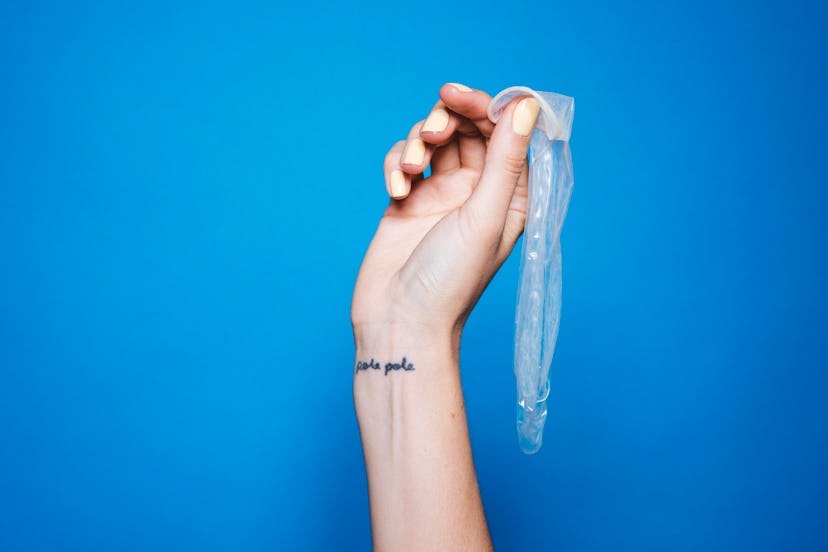 A female hand with a tattoo on a wrist holding a used latex condom in front of a blue background
