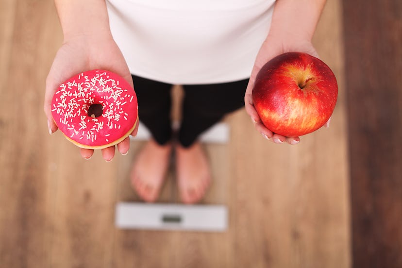 A woman standing on a scale and holding a donut in one, and an apple in her other hand