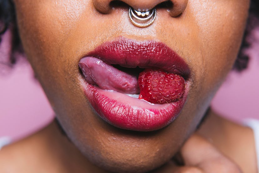 A woman with a nose ring wears red lipstick while sticking her tongue out and holding a raspberry in...